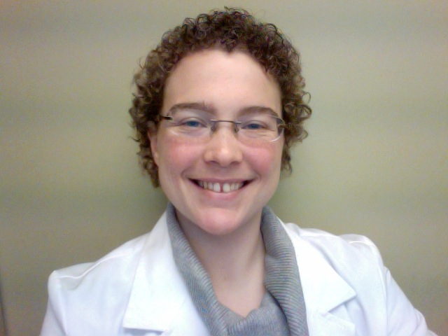 Brewster has short, curly, blonde hair and glasses, wearing a white lab coat.